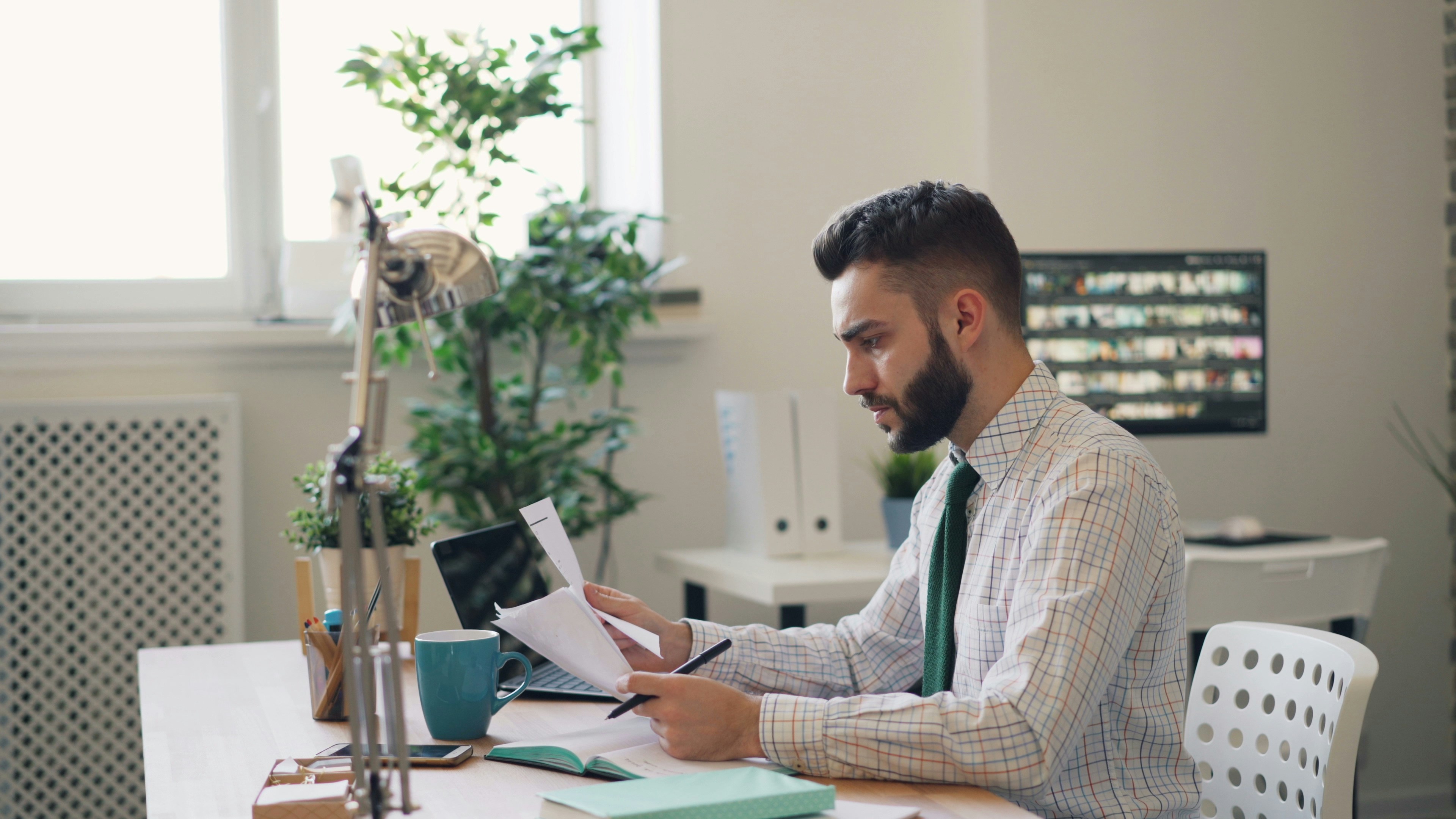 A businessman in a checkered shirt and tie sits at a desk, reviewing documents with a focused expression. His workspace includes a laptop, notebook, and office supplies, with greenery and a window in the background.
